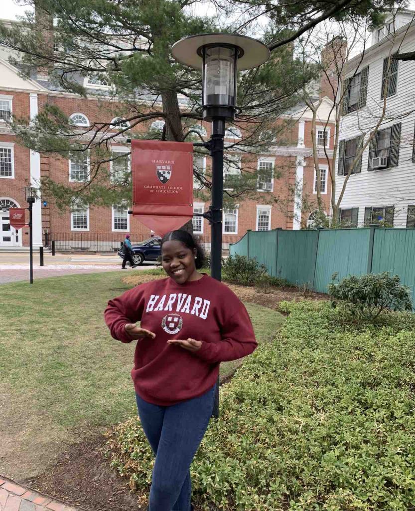 Moore stands in front of a green space on the Harvard campus wearing a crimson Harvard sweatshirt.